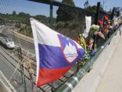 Ramos de flores y una bandera de Eslovenia recuerdan a las v&iacute;ctimas del accidente ferroviario del 24 de julio del a&ntilde;o pasado. De fondo, un tren de velocidad alta circula por la curva de A Grandeira.