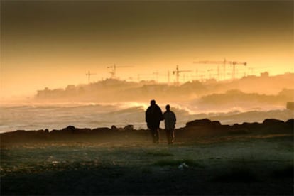 Una pareja pasea por una playa de Moncofa (Castellón) por delante de un fondo de grúas.