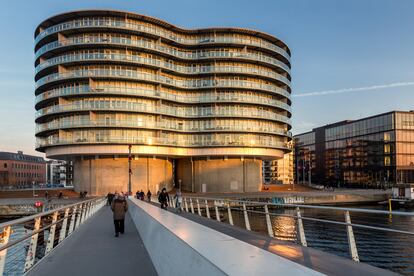 Frøsilo, viviendas ‘colgadas’ de antiguos silos (Copenhague, Dinamarca). Los apartamentos más deseados de Copenhague están construidos en dos antiguos silos cilíndricos de grano, pero no dentro —lo que hubiera condicionado su distribución e imposibilitado unas vistas fantásticas al mar— sino colgados de ellos mediante una funda o fachada de vidrio en forma de ocho, colocada sobre los volúmenes originales. En el espacio vacío de ambos silos de hormigón, cubierto con un techo de membrana plástica transparente, se disponen las tuberías y los conductos de instalaciones, los ascensores y las escaleras. Las 84 viviendas, de entre 90 y 200 metros cuadrados de superficie, se distribuyen en ocho plantas en la antigua zona del puerto de la capital danesa. El edificio se llama Frøsilo (silo, en danés), y ha sido diseñado por el estudio de arquitectura holandés MVRDV, que es el primero en calificar de “radical” su intervención. Fue reconocido como el mejor edificio de la ciudad en 2005, año de su inauguración.