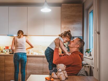 Photo of Busy Family Home With Father holding baby boy As Mother Prepares Meal during the day. Short hair mature father playing with son while sitting on kitchen table with mother working in background. Caucasian family in their kitchen, dad lifting baby in the air, mum cooking. Teddy Bear on table sitting.