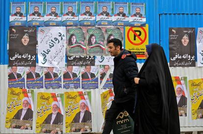 Una pareja iraní camina junto a  una pared con propaganda electoral.