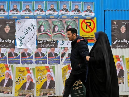 Una pareja iraní camina junto a  una pared con propaganda electoral.