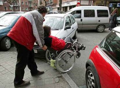 Una discapacitada y su acompañante avanzan entre coches mal aparcados en Madrid.