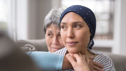 An ethnic woman wearing a headscarf and fighting cancer sits on the couch with her mother. She is in the foreground and her mom is behind her, with her arm wrapped around in an embrace, and they're both looking out the window in a quiet moment of contemplation.