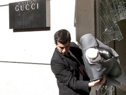Dos empleados de la tienda retiran trajes del escaparate roto.