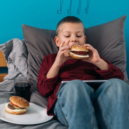 lazy young boy relaxing on bed in his room and using digital tablet and eating burgers junk food