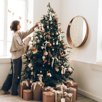 Young woman is decorating Christmas tree with many different decorations and festive garland in minimalist light interior. New Year celebration concept