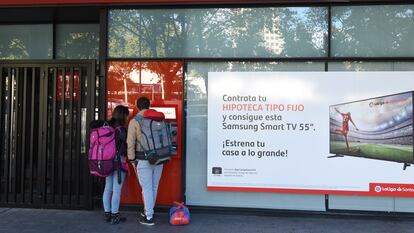 Dos personas retiran dinero de un cajero automático junto a un anuncio de hipotecas del Banco Santander, en una oficina de Madrid.