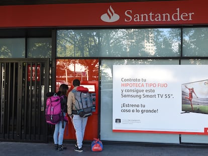 Dos personas retiran dinero de un cajero automático junto a un anuncio de hipotecas del Banco Santander, en una oficina de Madrid.