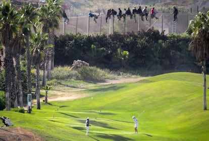 Una docena de inmigrantes permanece encaramado a la valla de Melilla mientras dos personas juegan al golf.