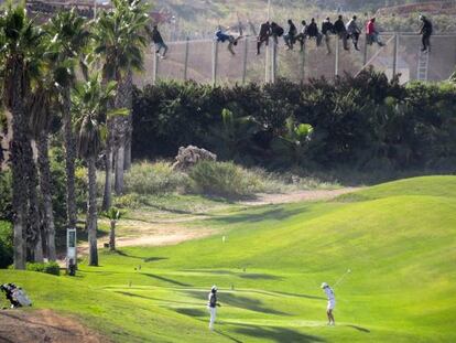 Una docena de inmigrantes permanece encaramado a la valla de Melilla mientras dos personas juegan al golf.