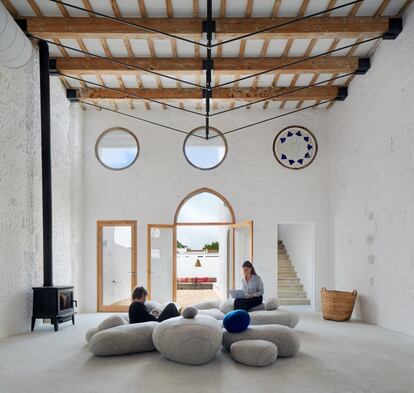 Una colección de cojines de lana Livingstones (producidos por Smarin) diseñados por la francesa Stephanie Marin amuebla el salón de la casa.<br><br>Esta es una casa colectiva, un espacio doméstico donde también se puede trabajar. Está pensado como lugar para vivir y teletrabajar cerca de la naturaleza. Y diseñado para un uso cambiante. Su dueña, la arquitecta Anna Truyol, pasó los veranos de su infancia en Menorca. Relacionaba el lugar con la tranquilidad. Y quiso trasladar esa idea al edificio. Fue su socia, Emma Martí, quien la convenció para que ese descanso fuera también visual. Juntas decidieron aprovechar la arquitectura popular mediterránea centenaria y añadirle un interior japonés, desnudo pero cómodo. Eso es este proyecto. En Sant Lluís, en el extremo suroriental de la isla, las arquitectas limpiaron el edifico de las sucesivas intervenciones que había sufrido y lo dejaron con su forma original. Lo desnudaron, deshaciendo tabiques, y pintaron la piedra local de marés, con sus imperfecciones, de blanco. Hoy la planta baja es una cocina con salida al patio. Las mesas se pueden unir o separar. Y hay una pizarra-pared para pensar, jugar o planificar. En la planta superior, el estudio Archimboldi que lidera Truyol ideó cubos-dormitorio fabricados con pino de Flandes. Los amuebló con futones de algodón y los cerró, como alcobas, con cortinas de lino. En el exterior, la piscina conforma un microclima con comedor y zona de estar construida con bancos corridos y pino de Flandes tratado para resistir la intemperie. El resultado es una vivienda sencilla, serena, actual y atemporal a la vez. <br>