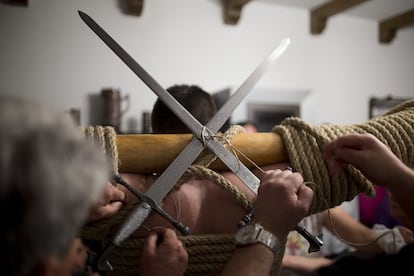 De cada uno de los brazos se cuelgan unos grandes eslabones de acero bien equilibrados y en la espalda se colocan dos espadas cruzadas apuntando al cielo.