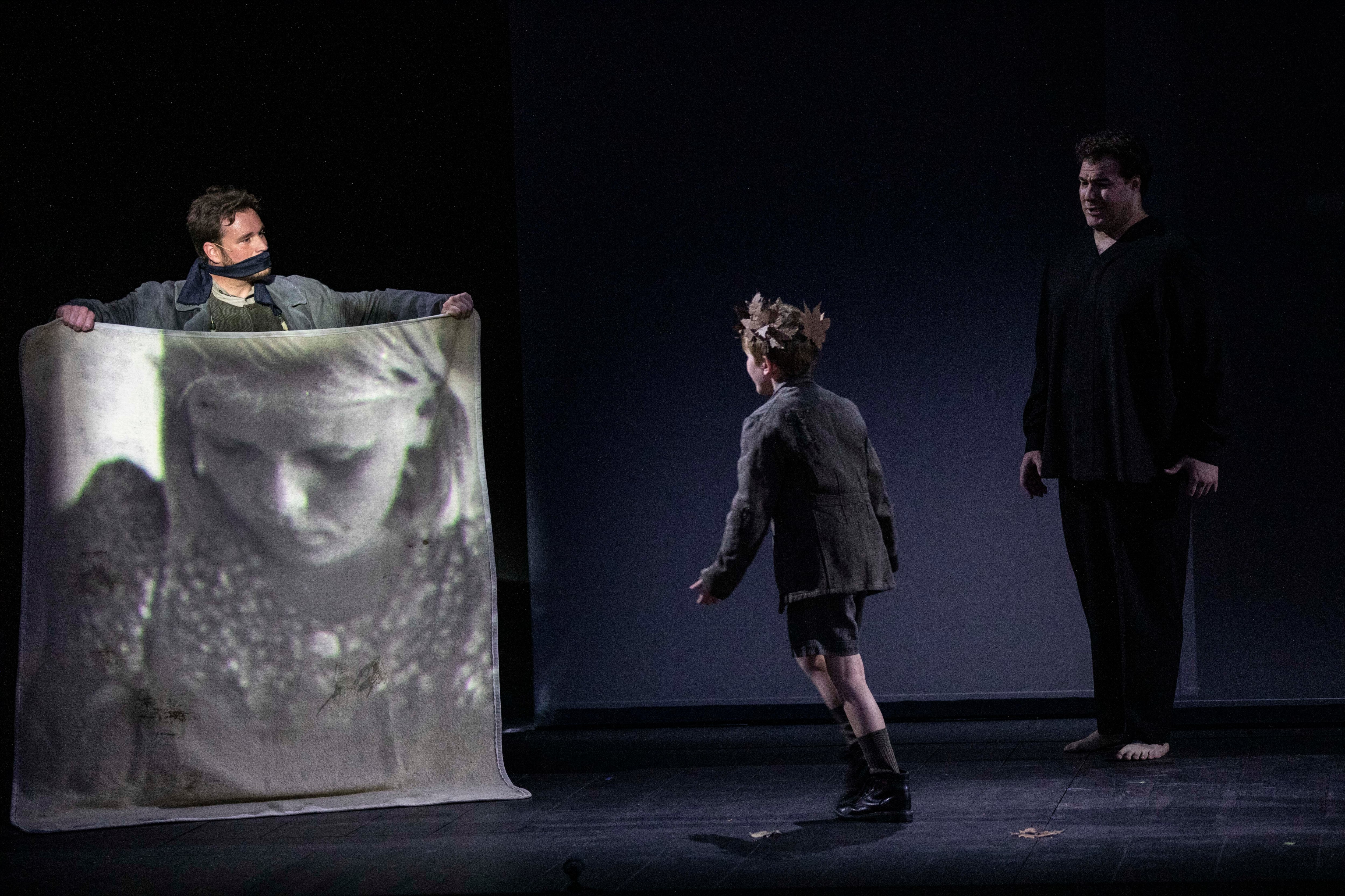 Papageno (con la boca vendada) muestra el retrato de Pamina al niño Tamino mientras canta el Tamino adulto (Mauro Peter) su aria ‘Dies Bildnis ist bezaubernd schön’.
