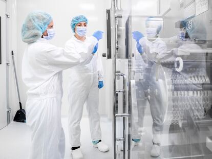 Dos mujeres con ropa de trabajo de seguridad vistas en un laboratorio farmacéutico frente a la compleja máquina que forma parte de la fabricación de medicamentos.