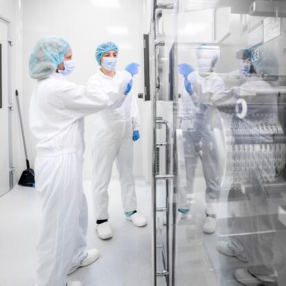 Dos mujeres con ropa de trabajo de seguridad vistas en un laboratorio farmacéutico frente a la compleja máquina que forma parte de la fabricación de medicamentos.