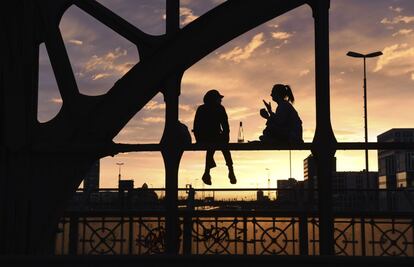 Dos mujeres silueteadas por la puesta de sol charlan sentadas sobre un puente en Munich (Alemania).