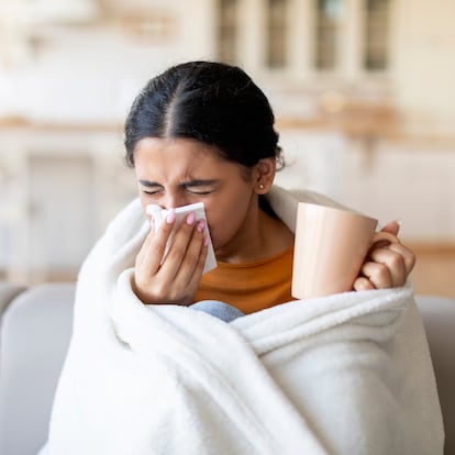 Cold And Flu Symptoms . Sick young indian woman covered in blanket blowing her running nose in a tissue while sitting on couch in living room, ill lady drinking hot tea, feeling unwell at home