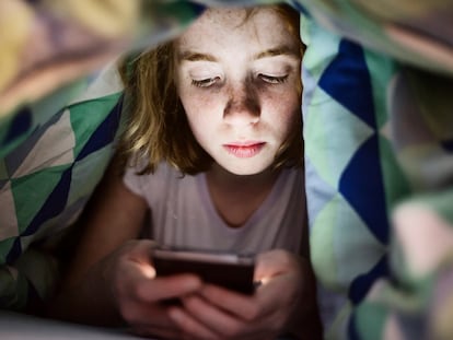 Una niña mira un teléfono móvil debajo de las sábanas.
