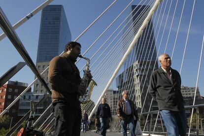 Un músico toca el saxofón en el puente de Zubizuri, en Bilbao, mientras varios ciudadanos atraviesan el monumento diseñado por el arquitecto e ingeniero Santiago Calatrava.