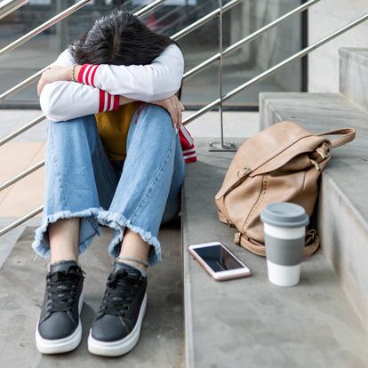 Full body of female student in casual outfit sitting on stairway with smartphone cup and backpack while having problem with bullying