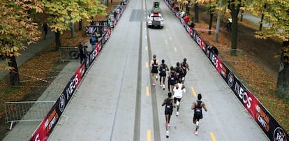 Kipchoge, de blanco, escoltado por un coche, las libres (en V) y dos corredores por detrás.