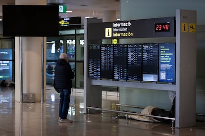 Una personas sin hogar duerme bajo un plafón informativo de la Terminal 1 mientras un usuario del aeropuerto consulta los horarios de los vuelos, este martes.