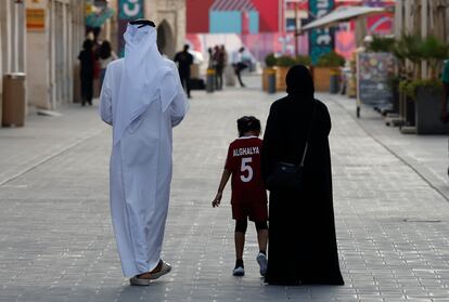 Un hombre vestido con el tradicional thobe árabe, una mujer de abaya negra hasta los pies y una niña con una camiseta de fútbol pasean por Doha.