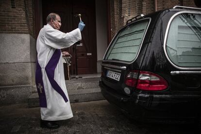 El diácono Santiago Pérez salpica agua bendita sobre el coche fúnebre de una difunta, tras oficiar un responso a la entrada de la capilla de La Almudena, Madrid, el 31 de marzo. 