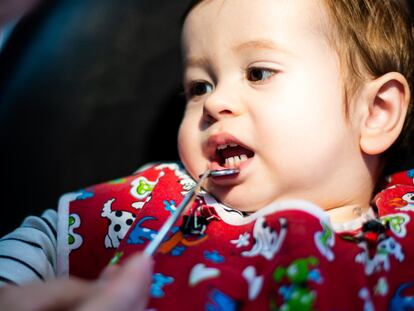 Un niño de menos de dos años en la consulta del dentista, con una bata roja, mira al dentista mientras este le baja el labio inferior con un pequeño espejo.