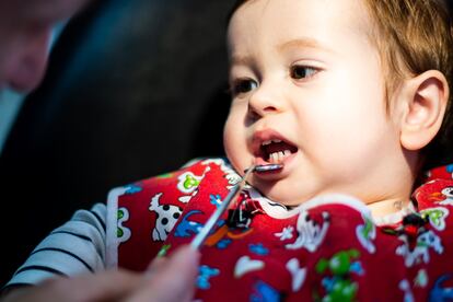 Un niño de menos de dos años en la consulta del dentista, con una bata roja, mira al dentista mientras este le baja el labio inferior con un pequeño espejo.