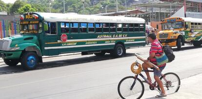 Un hombre circula en bicicleta por la avenida principal de La Libertad, cargando una manguera, tablones de madera y la tradicional ‘cebadera’ salvadoreña. En el fondo, los pintorescos buses de pasajeros de mediana y corta distancia.