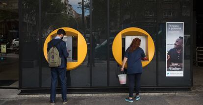 Una señora limpia un cajero de CaixaBank mientras un chico realiza una operación en la calle Orense (Madrid)