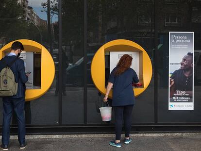 Una señora limpia un cajero de CaixaBank mientras un chico realiza una operación en la calle Orense (Madrid)