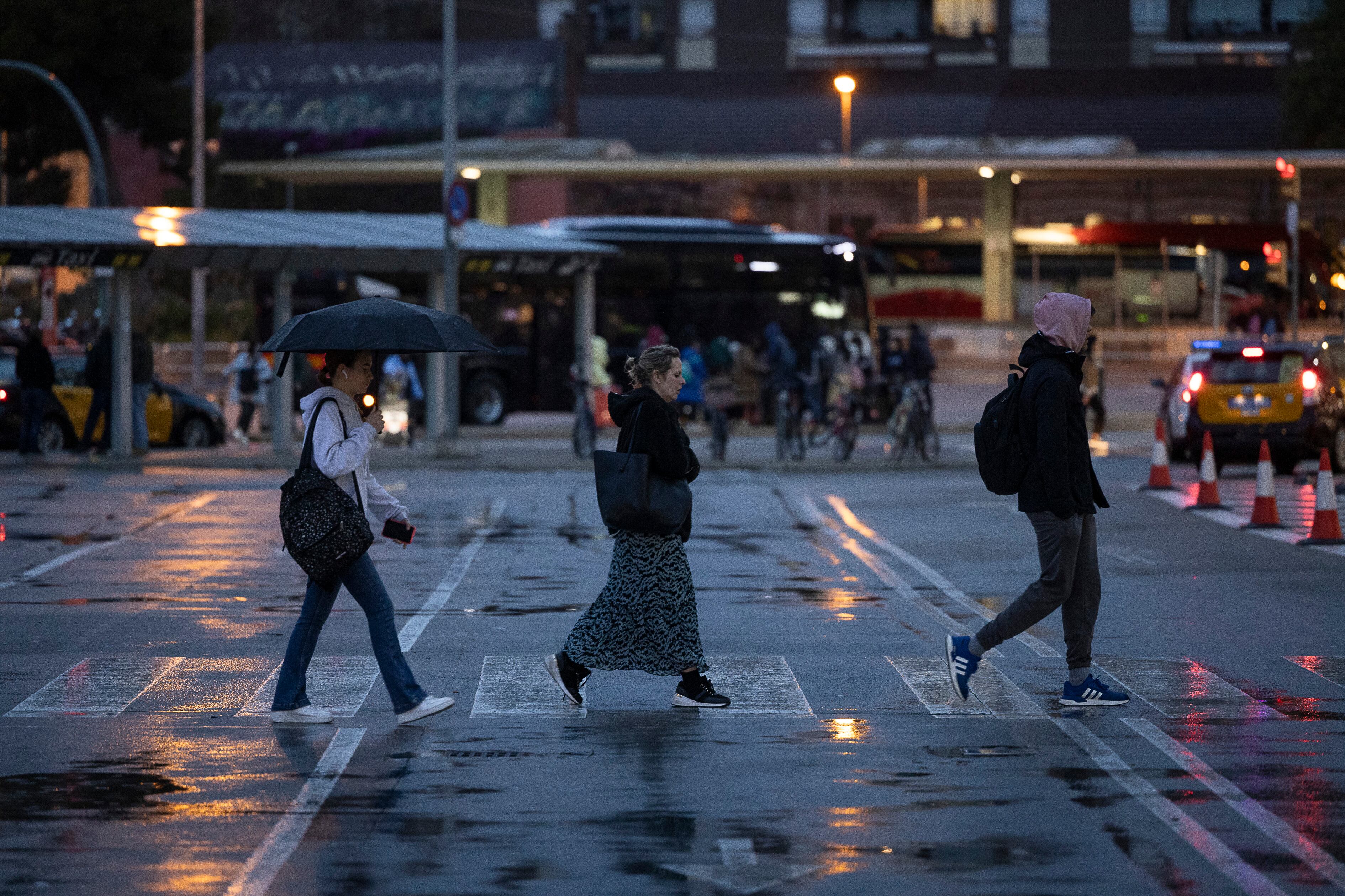 Tres personas cruzan un paso de cebra bajo la lluvia, en los exteriores de la estación de Sants, esta mañana en Barcelona.