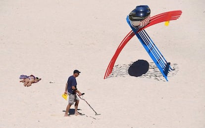 Una mujer toma el sol mientras un hombre con un detector de metales pasa delante de una de las esculturas que se exhiben, en la playa de Tamarama de Sídney.