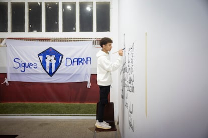 Zakaria Ezzouyn, pintando un mural con palabras elegidas por los futbolistas del FC Darna, el martes en la sala de la exposición 'Goals for Change'. A la izquierda, la bandera del club, diseñada por él.