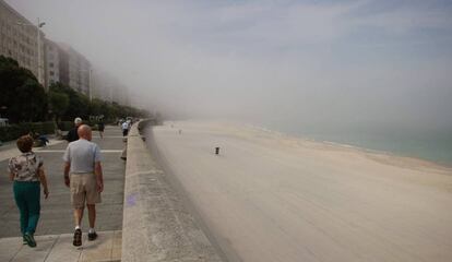 Varias personas caminan hoy por el paseo de la playa del Orzán de A Coruña cubierta por la niebla y sin bañistas.
