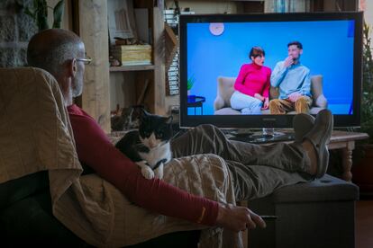 Un hombre, con el mando de la televisión en la mano y un gato en el regazo, mira una pantalla de televisión en su casa.

NOTA: El hombre es el fotógrafo Santi Burgos. Dvd 990 23.02.20 Un hombre ve la televisión en su casa. foto: Santi Burgos