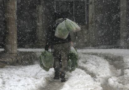 Un hombre cargado con bolsas de verdura camina bajo la nieve por una calle del barrio de Duma en Damasco, Siria.