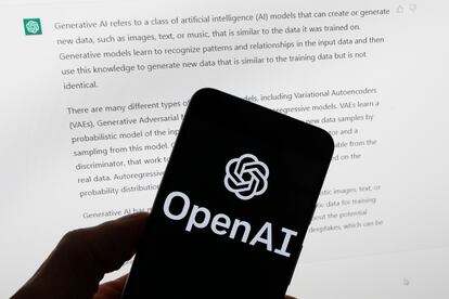 The OpenAI logo is seen on a mobile phone in front of a computer screen displaying output from ChatGPT, March 21, 2023, in Boston