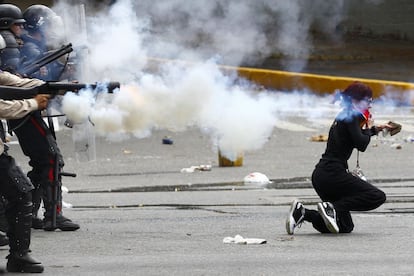 Uma manifestante se ajoelha segurando uma pedra enquanto membros da polícia Nacional venezuelana dispara gases lacrimogêneos.