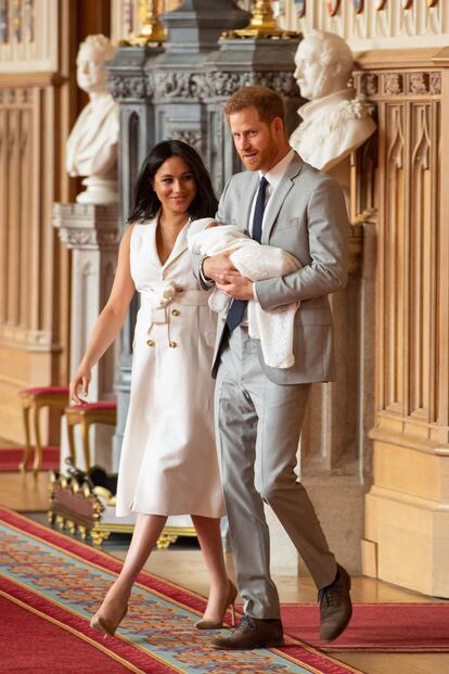 Para a apresentação de seu filho, Markle escolheu um vestido branco de botões, com um laço na cintura, que marca a silhueta. Harry, com seu filho nos braços, optou por um terno cinza claro uma gravata azul marinho