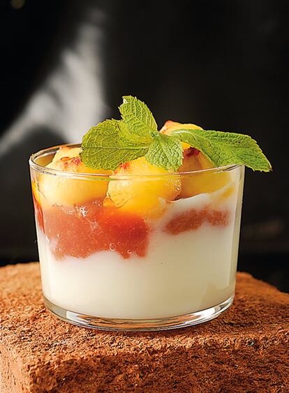 <b>Para 8 personas: Panna cotta: ½ litro de leche, ½ litro de nata, 6 cucharadas de azúcar, 2 sobres de preparado para hacer cuajada. Membrillo: 200 gramos de dulce de membrillo, 1 vaso de vino de agua. Fruta: 2 melocotones de viña, menta fresca</b> 1. Membrillo: Mezclar en un cazo el membrillo cortado en trocitos con el agua, acercarlo al fuego, dar unos hervores para que se licue y dejarlo enfriar. 2. <i>Pannacotta</i>: Separar un vaso de leche fría. Disolver el azúcar en el resto de leche con la nata y hacer hervir. 3. Disolver los polvos de cuajada en la leche fría. Añadir los polvos disueltos a la leche-nata cuando rompa a hervir, mover, dar un hervor y llenar vasos o copas hasta la mitad.  Cuando esté sólido, verter encima el membrillo y dejar que se solidifique. 4. <b>Presentación</b>: Media hora antes de servir, pelar y cortar en bolitas los melocotones, poner las bolas mezcladas con la menta unos minutos y cubrir las copas. 5. <b>Elaboración</b>: Preparar las copas completas, taparlas con papel film y guardar en la nevera o en el congelador. 6. <b>Conservación</b>: 2 días en la nevera, 1 mes en el congelador. 7. <b>Regeneración</b>: Sacar de la nevera o descongelar en la nevera y después dejar ½ hora a temperatura ambiente y cubrir con las bolas de melocotón y menta al momento de servir. 8. <b>Nota</b>: Se puede preparar una ensaladera o copa grande de cristal en lugar de copas individuales.