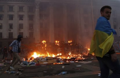 Un manifestante envuelto en una bandera ucrania camina frente al edificio de los sindicatos en llamas.