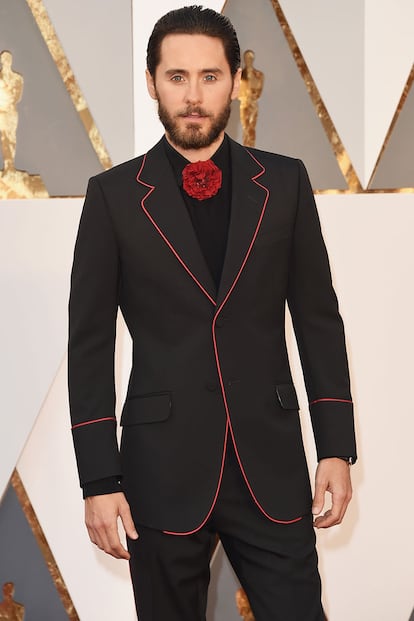 Jared Leto innovando en la alfombra roja con traje negro ribeteado en rojo y flor en lugar de pajarita. Todo de Gucci.