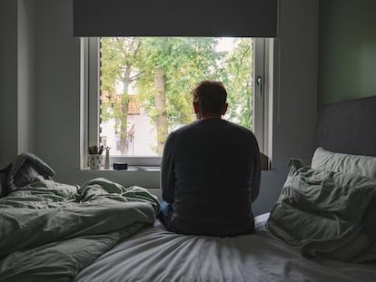 Un hombre mira por la ventana, sentado en kla cama, desde su habitación.