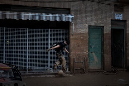 Un joven juega con una pelota embarrada en la acera de la calle San Juan de Ribera, Paiporta, mientras los trabajos de limpieza continúan tras la riada. Voluntarios y fuerzas de seguridad vacían portales llenos de barro, y una excavadora manejada por un joven voluntario de Castilla-La Mancha carga el lodo en un camión. El 11 de noviembre era mi primer día en Paiporta y recorría la ciudad sin saber del todo qué me iba a encontrar. Al acercarme a la parroquia de San Ramón, el nivel de barro en las calles iba en aumento. Al girar una esquina, el movimiento en un portal llamó mi atención: hombres sacaban escombros mientras en medio de la escena, un chico comenzó a patear una pelota cubierta de barro. En ese instante, recordé las palabras de Pelé: "El fútbol es música, baile y armonía. Y no hay nada más alegre que la esfera que rebota."