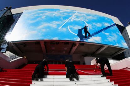 Tres operarios instalan la alfombra roja del Festival de Cannes a la entrada del Palacio de Festivales, decorado con el póster del certamen, que homenajea a 'El show de Truman'.