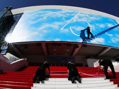 Tres operarios instalan la alfombra roja del Festival de Cannes a la entrada del Palacio de Festivales, decorado con el póster del certamen, que homenajea a 'El show de Truman'.